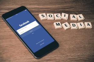 A smart phone lying on a wooden table displaying Facebook next to a collection of scrabble pieces that spell out social media.