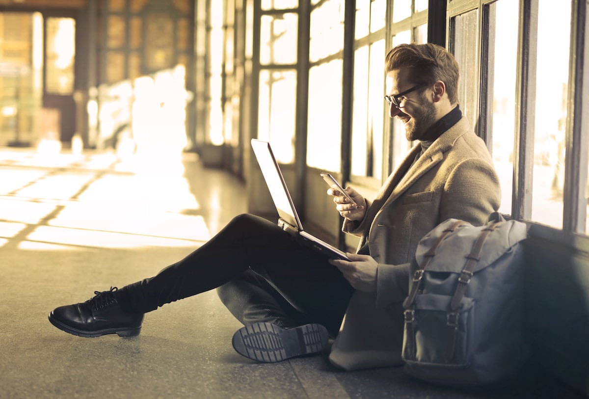 A man sitting on the ground with his smart phone and his laptop in front of him, smiling at what he's seeing.